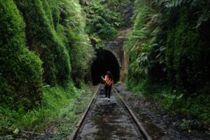A woman carrying a walking stick and a backpack heads through lush greenery towards a dark train tunnel.