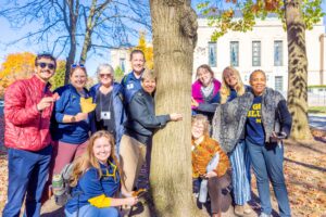 People smiling around a tree outside in the sun