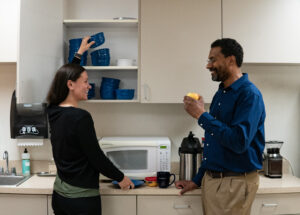 Two people are chatting and smiling in their workspace breakroom. One of them is reaching for a glass bowl from a cabinet full of reusable dishware. The other is eating an apple and using a reusable mug.