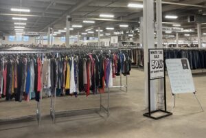 A rack of shirts is in the front of a big warehouse-style room. In the background, there are more rows of racks holding different kinds of clothes. A sign says that items are 50% off marked price.