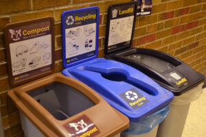 Photo of 3 U-M waste bins in order from left to right: compost, recycling, landfill. The bins are open and each has a sign listing what can be disposed in it.