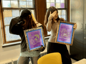 Two students hold screen printing frames up in front of a stained glass window. They are ducking their head under their hands.