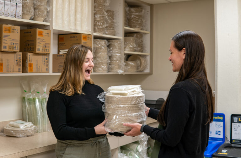 Two people hold a pile of compostable plates and napkins, with more kitchen serviceware in the background.