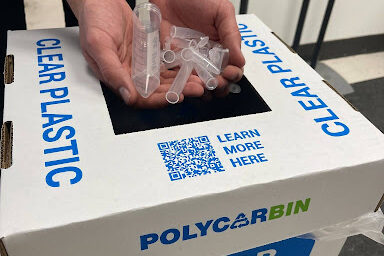 Someone holds pipette tips over a bin labeled "Polycarbin: Clear Plastic" for recycling.