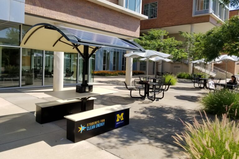Photo of an outdoors picnic table with two benches. Above the table, there are solar panels mounted to provide shade and power electric outlets.