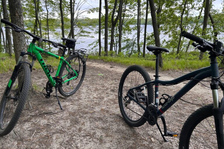 Two bikes are on a dirt trail surrounded by trees. There is a lake in the background.