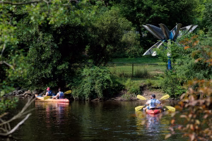 People in two kayaks paddle in the Huron River. Trees, grass, and a sculpture are in the background.